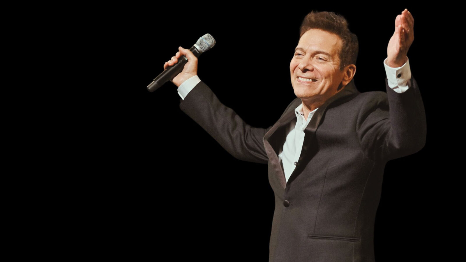 A man dressed in a dark suit and white shirt stands against a black background, holding a microphone in his right hand and extending his left arm outward in a welcoming gesture. He has a warm smile on his face, exuding confidence and charisma. His posture suggests he is engaging with an audience, possibly at the end of a performance or speech. The dark background focuses all attention on the man, highlighting his presence and expression.