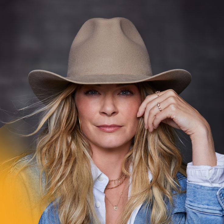 LeAnn with long, wavy blonde hair, wearing a wide-brimmed beige hat. She is dressed in a denim jacket over a white blouse. Her right hand is gently touching the brim of her hat, while her left hand rests near her face, showcasing a delicate ring on her finger. She gazes directly at the camera with a calm and confident expression. The background is dark and blurred, highlighting her as the focal point of the image.