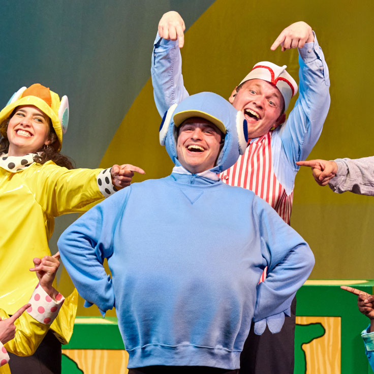 A group of performers is captured in a lively, joyful moment on stage. The central figure, dressed in a light blue costume with a helmet that has ear-like flaps, stands with a broad smile. To his right, a performer in a red and white striped outfit enthusiastically points at him while smiling widely. To his left, a woman wearing a yellow raincoat and a hat with animal ears also points at him, beaming with happiness. The colorful background and the characters' expressive poses and costumes create a fun and energetic atmosphere.