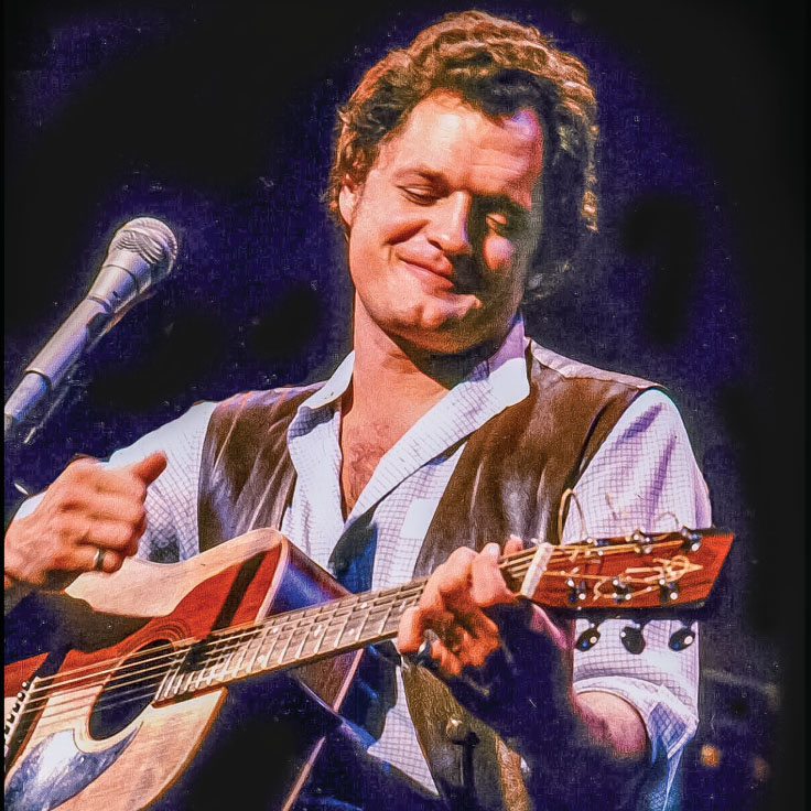 A man is performing on stage, playing an acoustic guitar and smiling warmly. He has curly hair and is wearing a white shirt with rolled-up sleeves and a leather vest. The background is dark, with stage lighting highlighting his face and guitar, giving a sense of an intimate, soulful performance. A microphone is visible to his left, indicating he is singing while playing. His expression and posture convey a deep connection to the music and the audience.