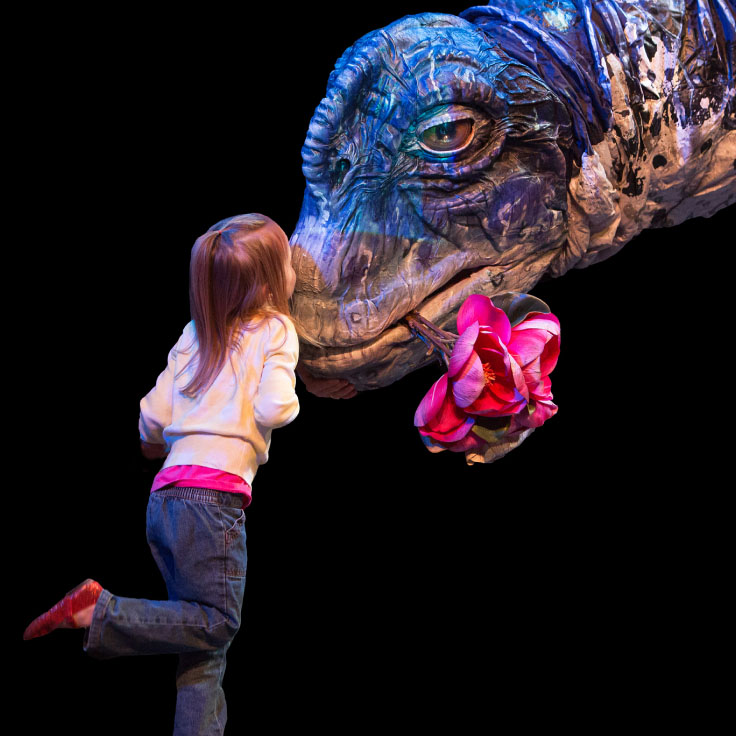 A young girl with long brown hair, wearing a white sweater and blue jeans, leans forward to touch noses with a large, realistic dinosaur puppet that looks to be a Sauropod. The dinosaur's head is intricately detailed with blue and grey textures, and it holds a pink flower in its mouth.