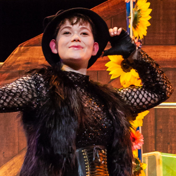 A performer, dressed in a black, gothic-style outfit, is captured in mid-pose. She wears a wide-brimmed black hat, a black netted top, and a fur vest. Her outfit is accentuated with a wide belt featuring metallic details. She stands confidently with a slight smile, exuding a sense of pride and charisma. Bright sunflowers are visible in the background, adding a vibrant contrast to her dark attire and enhancing the overall theatrical atmosphere.