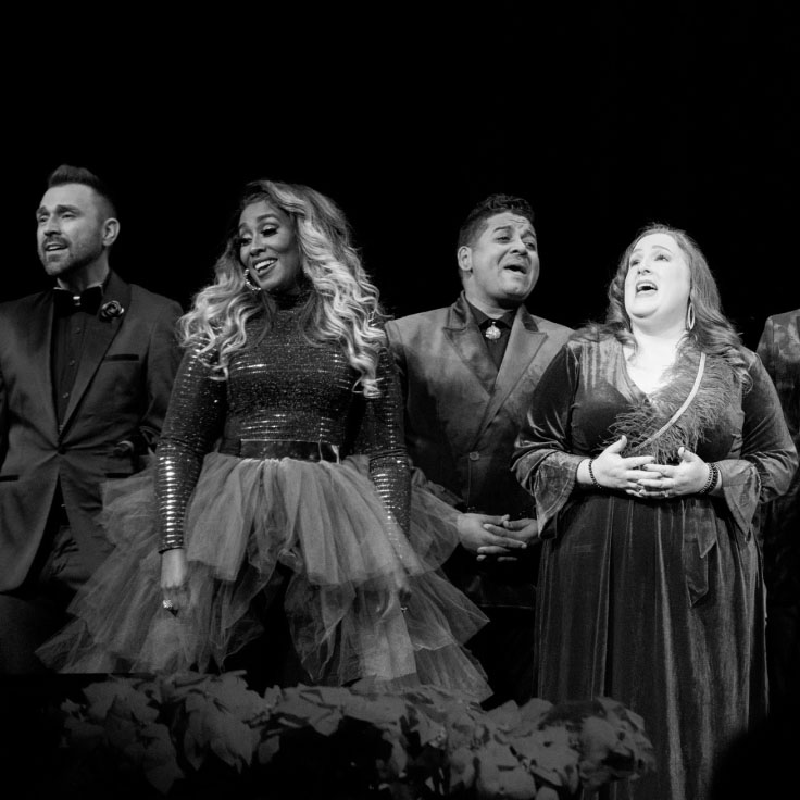 A group of 4 singers stands on stage, captured in a black and white photo. They are arranged in a line, with expressions of joy and passion as they perform. The women are dressed in elegant dresses, some with sequins and some with tulle, while the men wear sharp suits. The stage background is dark, emphasizing the performers who are brightly lit, creating a striking contrast. The audience is partially visible in the foreground, watching the performance attentively.