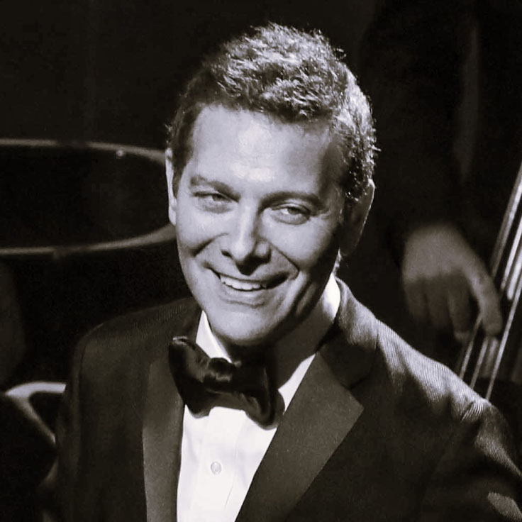 A black and white photo captures a man in a tuxedo with a bow tie, smiling warmly. He has neatly styled hair and a confident, charismatic demeanor. The background is dimly lit, likely indicating a performance setting, with a faint outline of a double bass and a piano, suggesting he is at a musical event. The lighting highlights his face, creating a classic and elegant atmosphere.