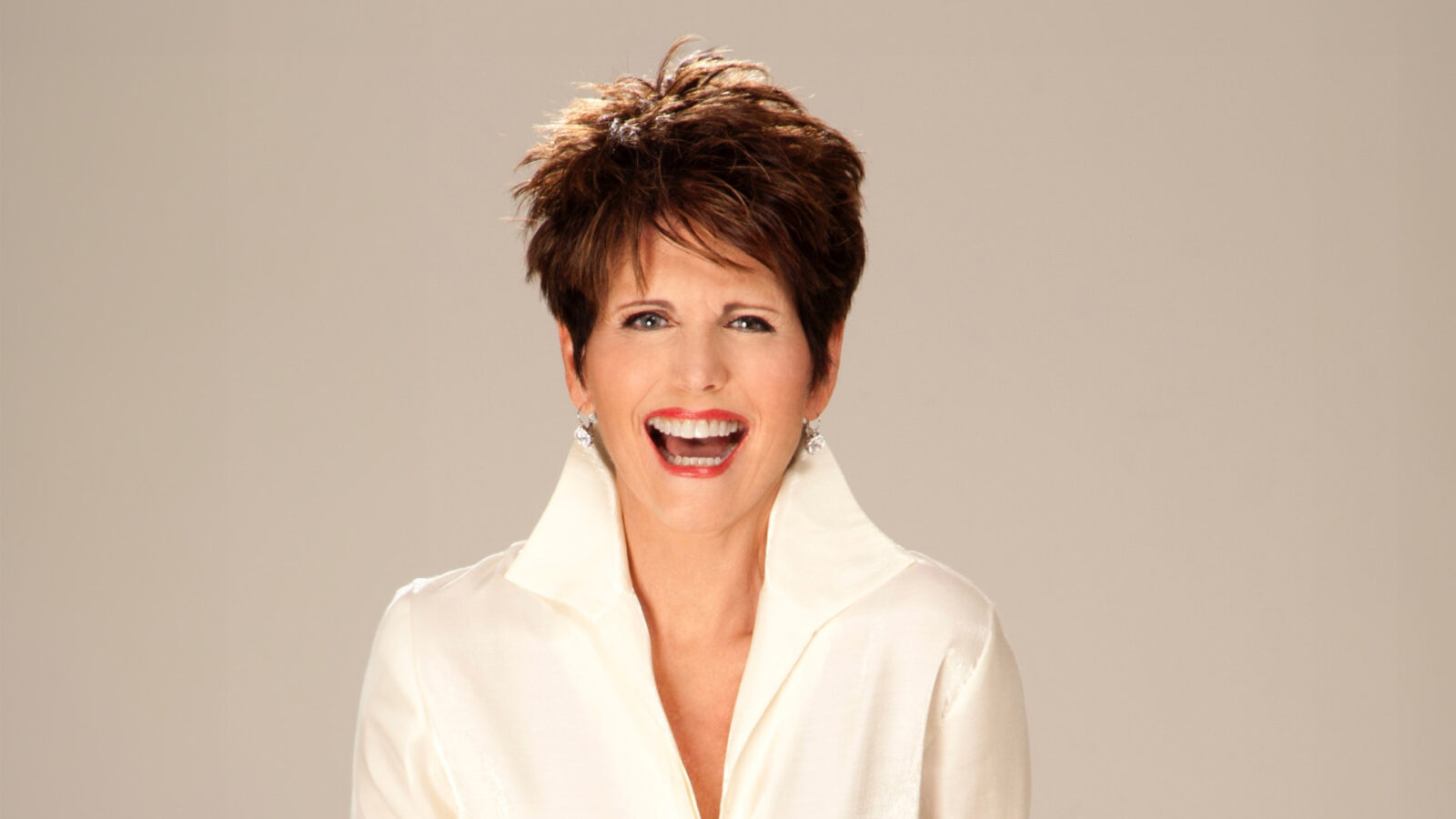 A woman with short, styled brown hair, smiling broadly and wearing a white collared shirt. She has a joyful expression and is accessorized with silver earrings.