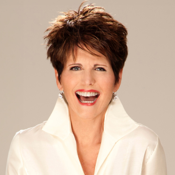 A woman with short, styled brown hair, smiling broadly and wearing a white collared shirt. She has a joyful expression and is accessorized with silver earrings.