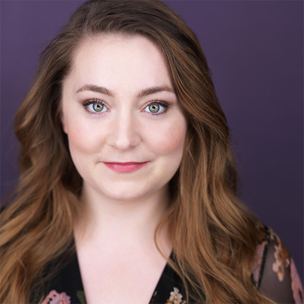 A professional headshot of a woman with long, wavy light brown hair. She has blue-green eyes and is wearing subtle makeup with a natural look. She is smiling softly, dressed in a floral-patterned top, and standing against a purple background.