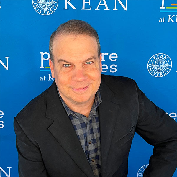 A headshot of a man with short, light brown hair, smiling at the camera. He is wearing a black blazer over a plaid shirt and is posed against a blue backdrop with Kean University logos