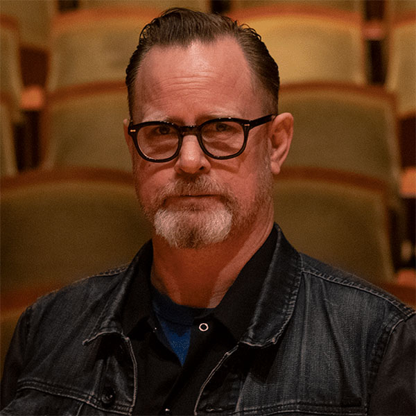 Headshot of a man wearing glasses and dark shirt with theatre seats as his background