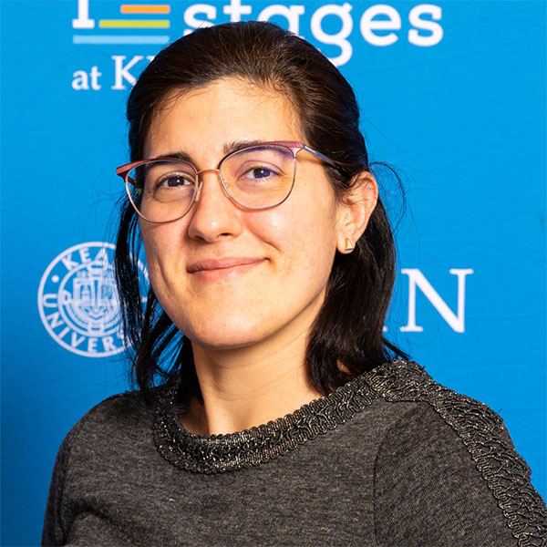 Headshot of a woman wearing glasses and smiling at the camera in a dark shirt and light blue background