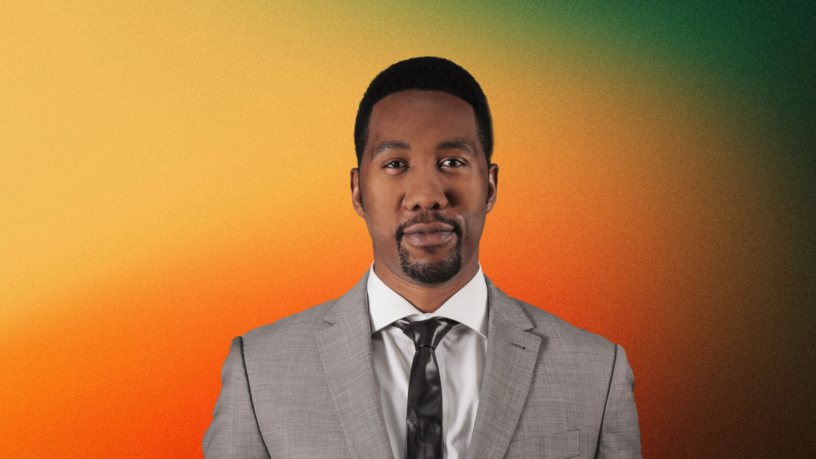 Portrait of Ndaba Mandela, grandson of Nelson Mandela, wearing a light grey suit, white shirt, and a dark tie. He has a short beard and mustache, and stands against a gradient background transitioning from orange to green.