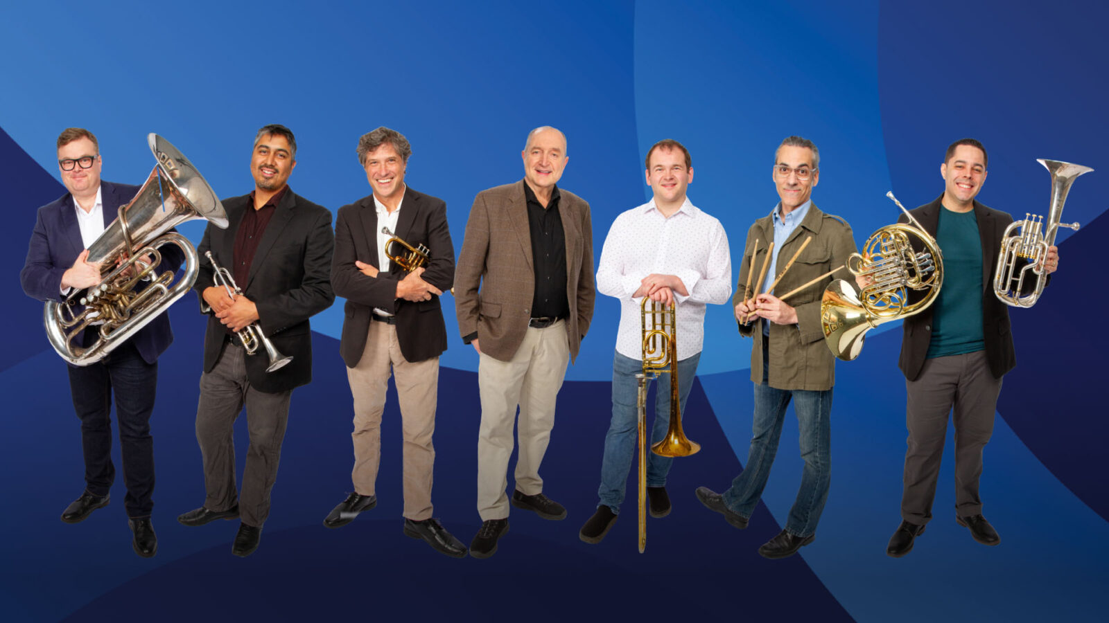 The Dallas Brass ensemble poses in a playful formation against a light beige background. Six musicians, dressed in black shirts and jeans, stand in a single file line, each holding their brass instrument in front of them. The instruments, including trumpets, trombone, tuba, and French horn, are aligned vertically from top to bottom, creating a visually striking effect. The musicians are smiling, exuding a sense of camaraderie and fun, with the member at the front kneeling and holding a large silver tuba. The arrangement highlights both the instruments and the group's unity.