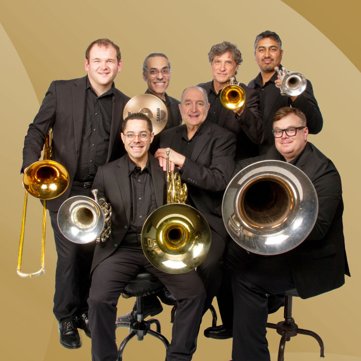 The Dallas Brass ensemble poses in a playful formation against a light beige background. Six musicians, dressed in black shirts and jeans, stand in a single file line, each holding their brass instrument in front of them. The instruments, including trumpets, trombone, tuba, and French horn, are aligned vertically from top to bottom, creating a visually striking effect. The musicians are smiling, exuding a sense of camaraderie and fun, with the member at the front kneeling and holding a large silver tuba. The arrangement highlights both the instruments and the group's unity.