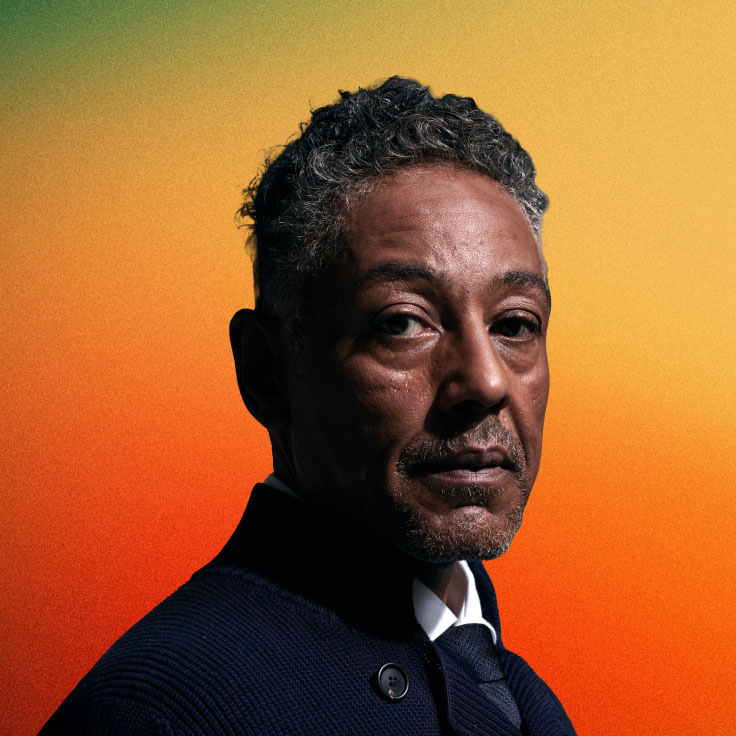 Portrait of Giancarlo Esposito, actor known for his roles in 'Breaking Bad' and 'The Mandalorian,' wearing a dark textured jacket over a white shirt. He has short, curly grey hair and a trimmed goatee, with a contemplative expression as he looks slightly off to the side. The background is a gradient transitioning from green to orange.