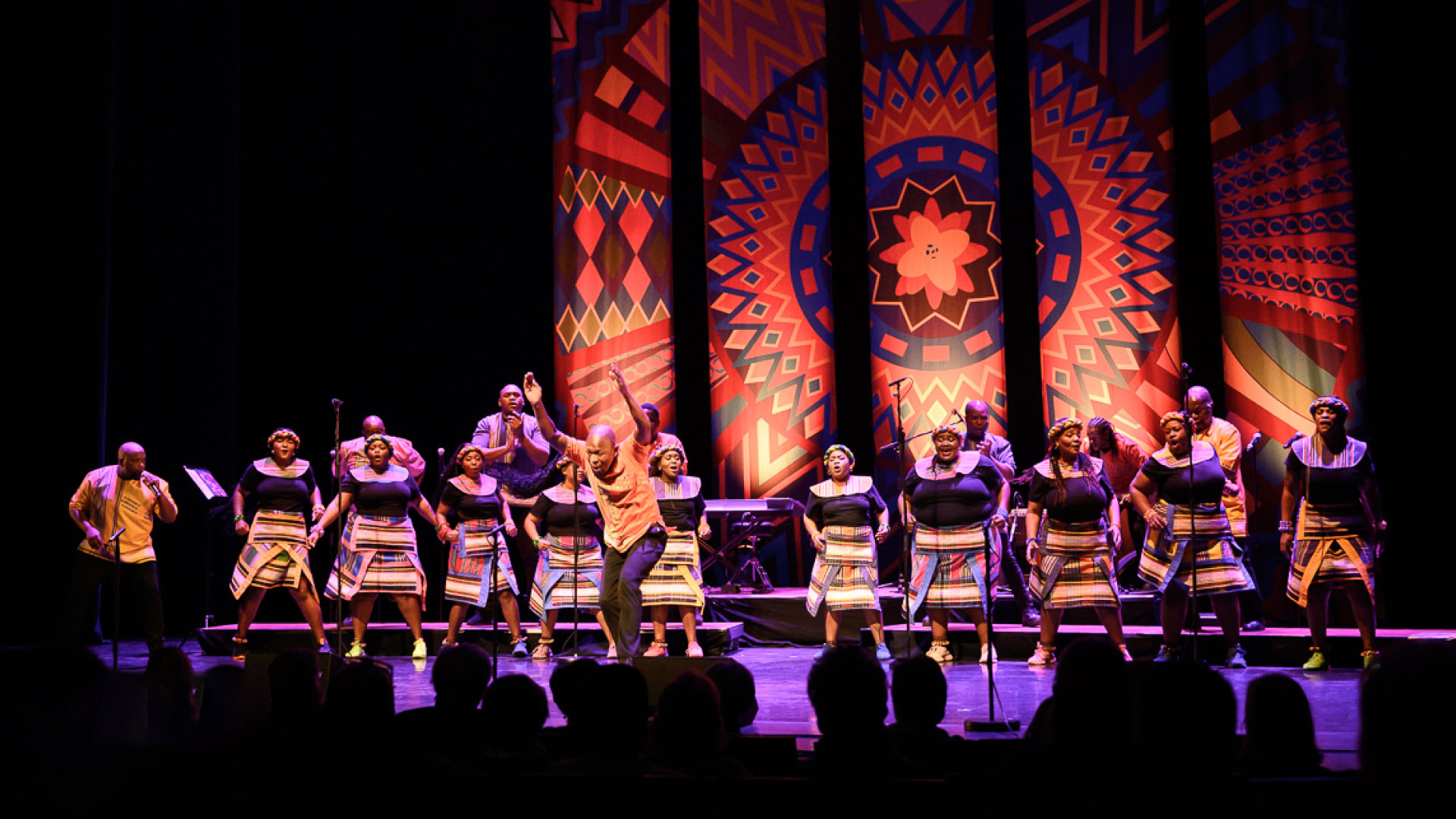 A group of performers clothed in colorful traditional attire engage in a dynamic performance on a stage adorned with vividly patterned backdrops.