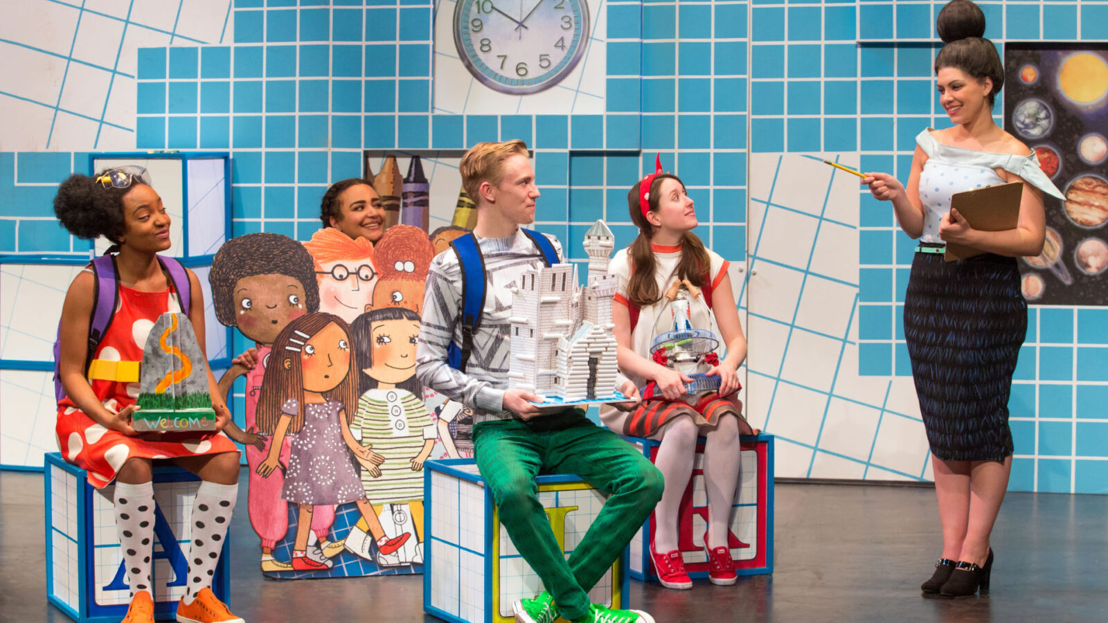 A lively classroom scene with four students showcasing their creative projects in front of a teacher. The setting features vivid, artistic backdrops and props. The teacher holds a clipboard, indicating an assessment or presentation activity.