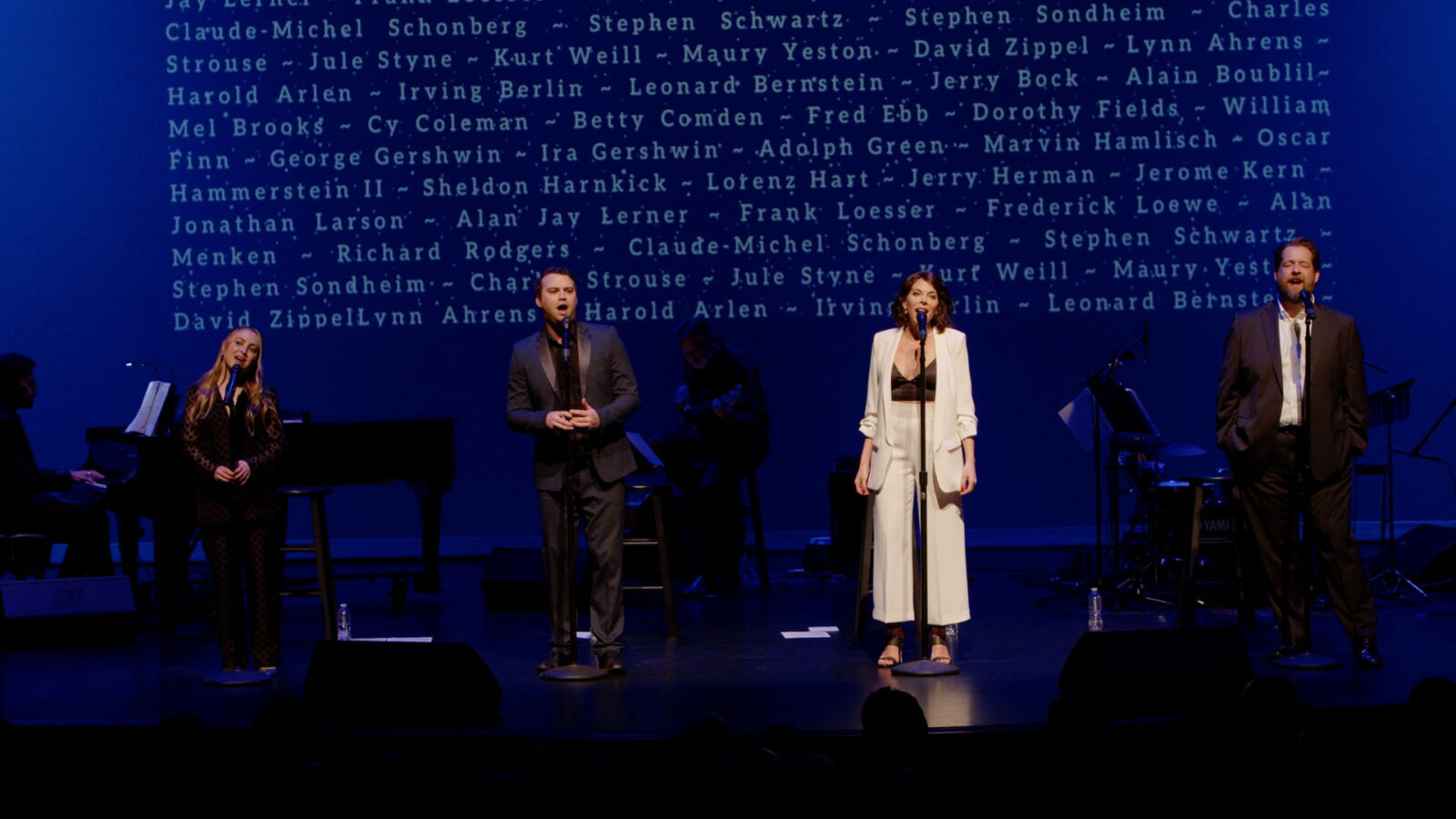 Performers stand on a dimly lit stage with visible names of renowned composers and lyricists projected behind them. Dressed in formal attire, they are engaged in a music recital, with musicians playing instruments to the left.