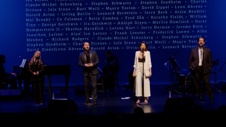 Performers stand on a dimly lit stage with visible names of renowned composers and lyricists projected behind them. Dressed in formal attire, they are engaged in a music recital, with musicians playing instruments to the left.