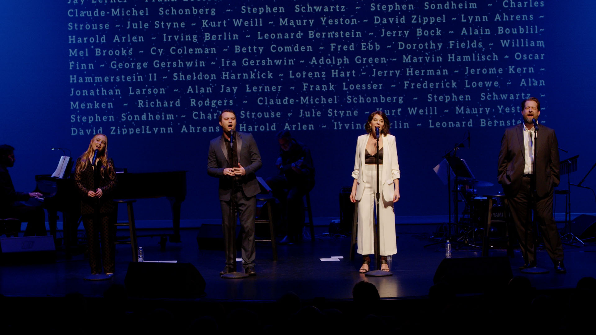 Performers stand on a dimly lit stage with visible names of renowned composers and lyricists projected behind them. Dressed in formal attire, they are engaged in a music recital, with musicians playing instruments to the left.