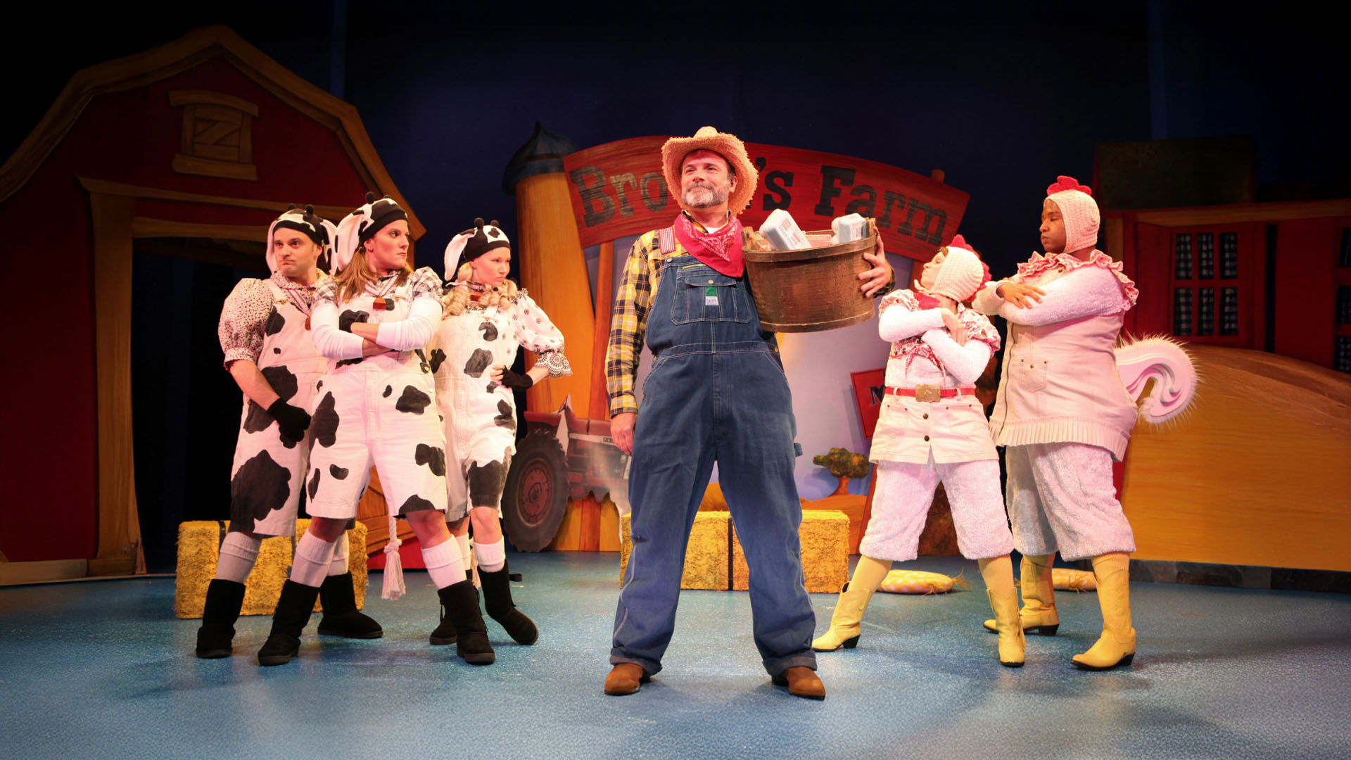 A theatrical production featuring characters dressed as farm animals alongside a farmer in a rustic setting. The stage is vibrant with colorful costumes, including cows and chickens, enhancing the whimsical farm ambiance. The farmer, dressed in traditional attire, holds a basket, suggesting a storyline of farm life and harvest. The background includes farm structures like barns and haystacks, creating an authentic rural atmosphere.