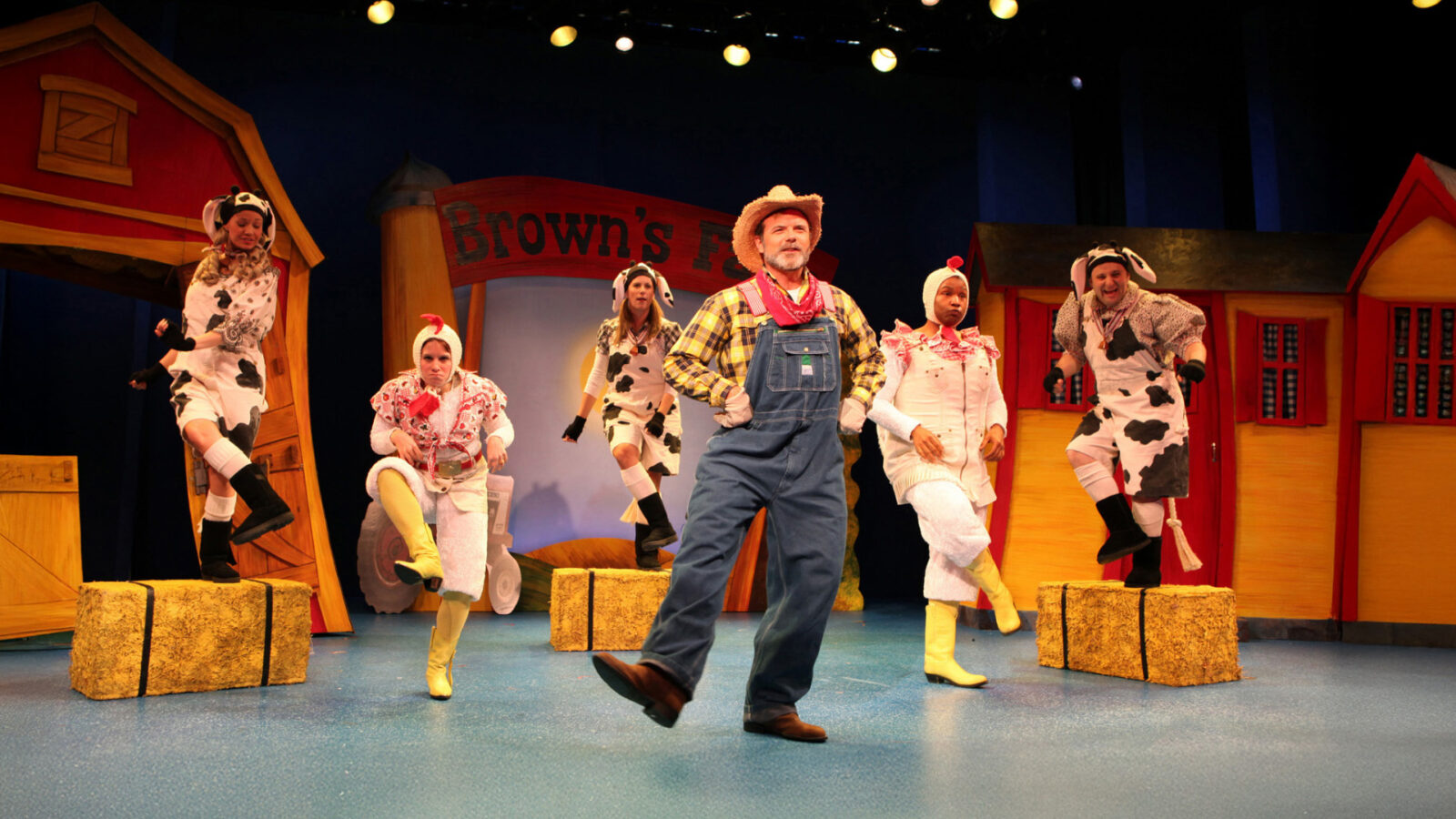 Performers in farm-themed costumes, including cows and a farmer, are energetically dancing on stage. The vibrant set features elements reminiscent of a barnyard, enhanced by colorful lighting that creates a lively and whimsical atmosphere.