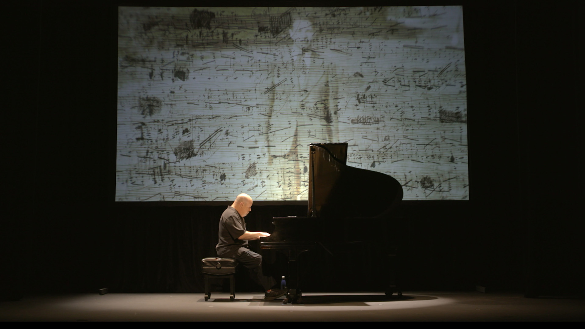 A solo pianist sits intently at a grand piano on a dimly lit stage. Above and behind the pianist, a large screen showcases an artistic visual featuring a blend of musical notes and a sketched figure.