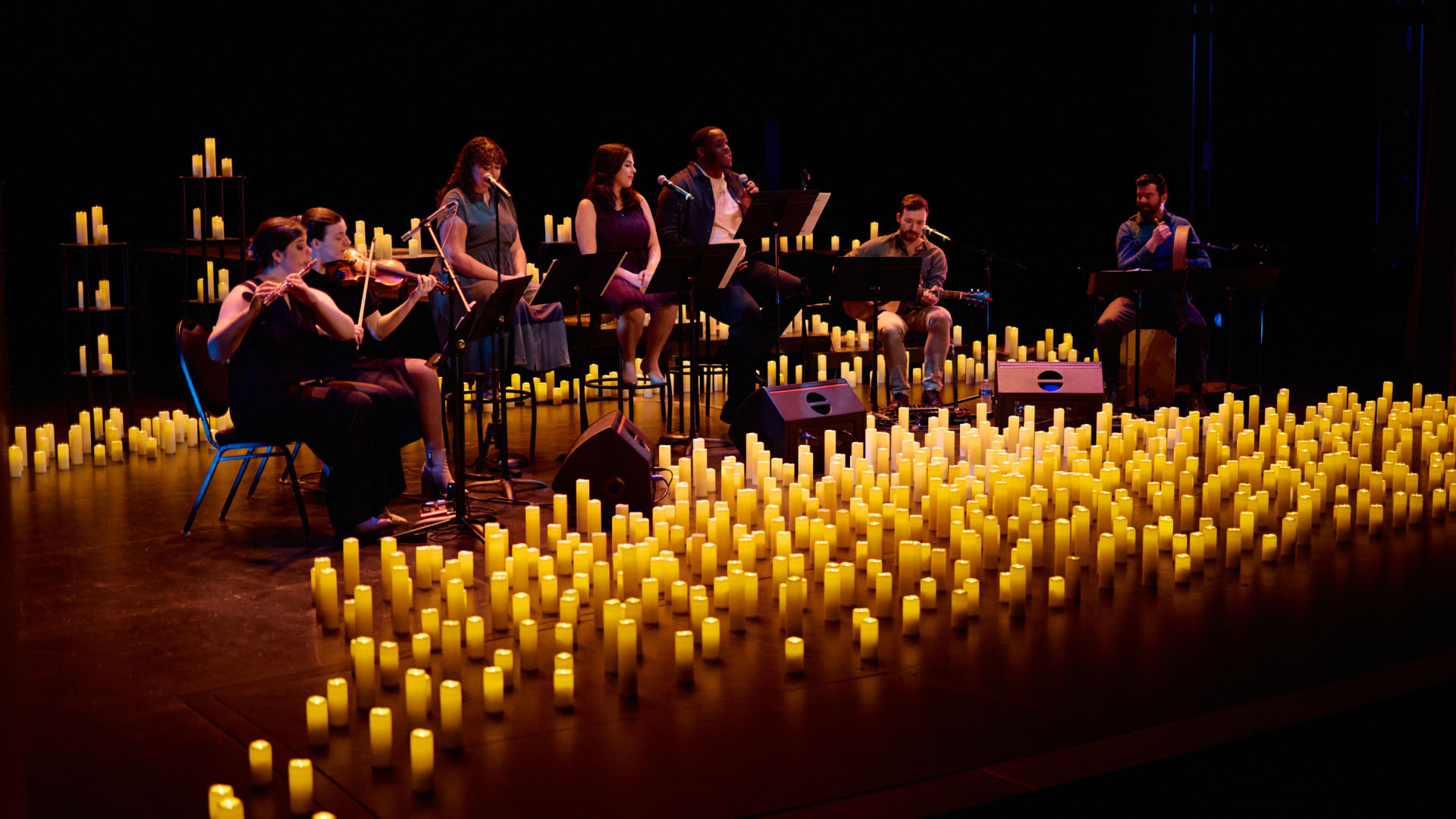 A group of musicians performs on a dimly lit stage surrounded by numerous yellow candles creating an enchanting and cozy atmosphere. The performers, playing various instruments including violins, are seated in a semi-circle.