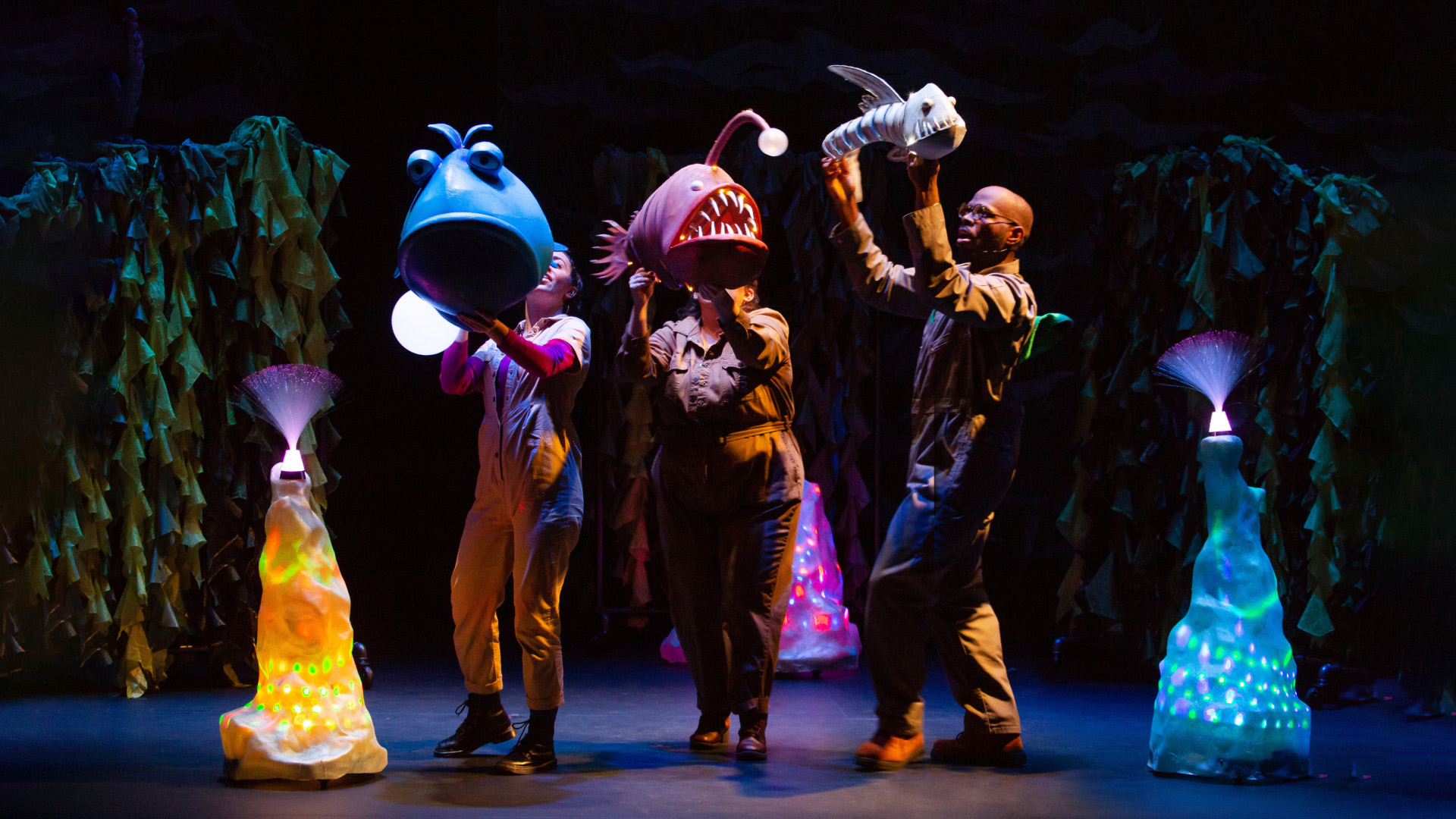 A stage performance featuring actors in vibrant, oversized fish costumes, illuminated under dark lighting to create an underwater scene. The production captures the imaginative portrayal of aquatic life, complete with colorful props and creative set design, conveying a sense of whimsy and fantasy typical of an underwater adventure.