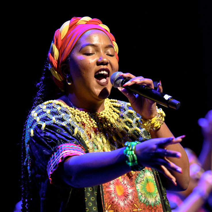 A performer passionately sings on stage, dressed in colorful traditional attire, including a patterned dress and a vibrant headwrap. The intricate beadwork and bright colors of the outfit reflect cultural heritage and celebration.