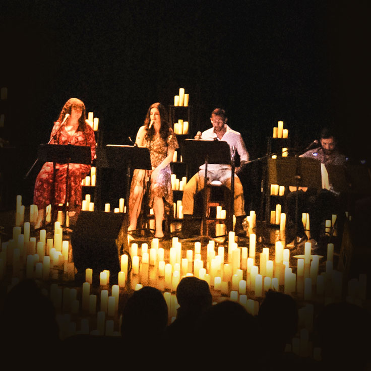 A dynamic musical ensemble performs on a dimly lit stage surrounded by numerous candles, creating an intimate and atmospheric setting. The warm glow of the candles enhances the live performance, highlighting the musicians and contributing to a serene and captivating ambiance. Audience members are silhouettes in the foreground, focused on the performance.