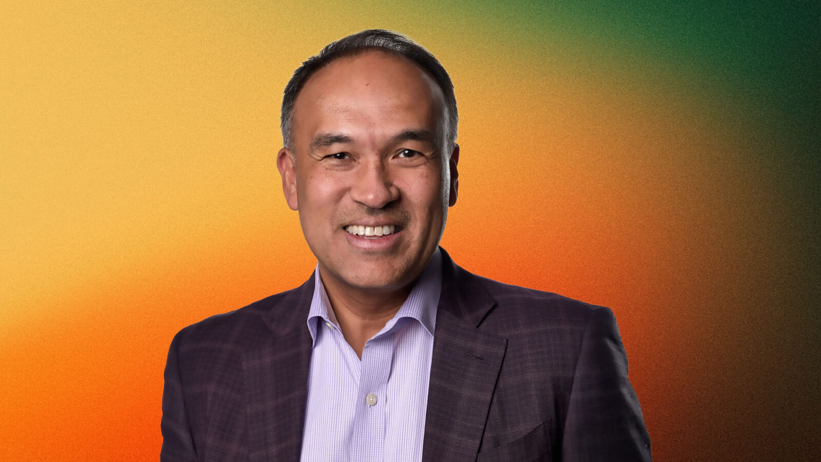 Mark Tatum with short dark hair and a broad smile, wearing a dark checkered blazer over a light purple shirt, against a warm orange and green gradient background.