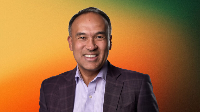 Mark Tatum with short dark hair and a broad smile, wearing a dark checkered blazer over a light purple shirt, against a warm orange and green gradient background.