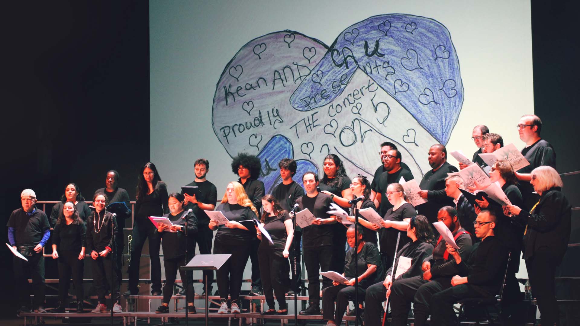 A diverse group of performers stands on stage, passionately engaging in a choir performance. They are dressed in black, emphasizing unity and professionalism. Behind them, a large, colorful drawing depicts hearts and motivating words, adding an artistic and inspiring backdrop to the performance.