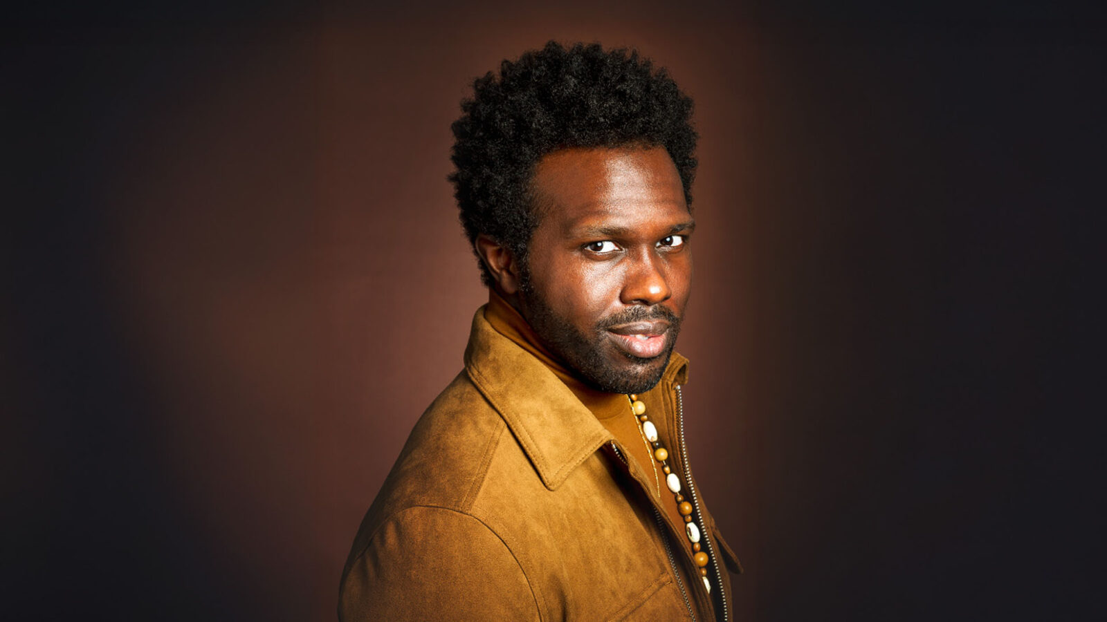 A contemplative man with curly hair is captured in a studio setting. He wears a brown suede jacket, complemented by a dark background that creates a warm and introspective atmosphere. His confident posture and direct gaze evoke a sense of depth and personality, highlighting a modern yet timeless fashion style.