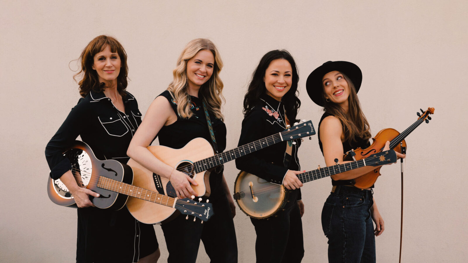 Four women musicians stand together, each holding a different string instrument including guitar, banjo, and violin. They are dressed in casual yet stylish attire, with a backdrop of a plain wall, suggesting a setting ready for a casual or intimate performance. Smiling and at ease, the group conveys a sense of camaraderie and joy in music-making.