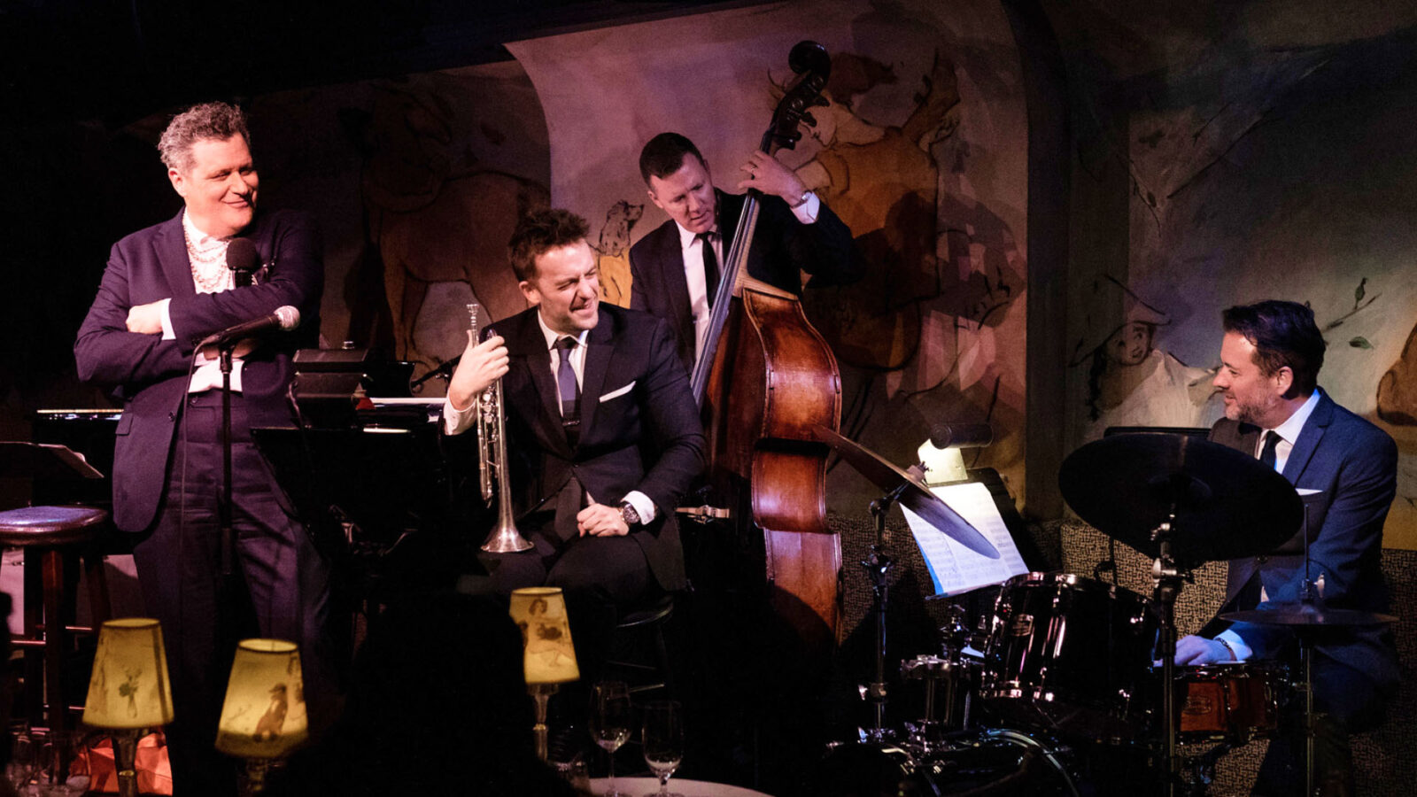 An intimate jazz club setting where a small ensemble of musicians performs. A trumpet player and a drummer are engaged in the music, while a stand-up bassist adds rhythm in the background. The atmosphere is warm and lively, enhanced by dim lighting and a vintage mural on the wall.