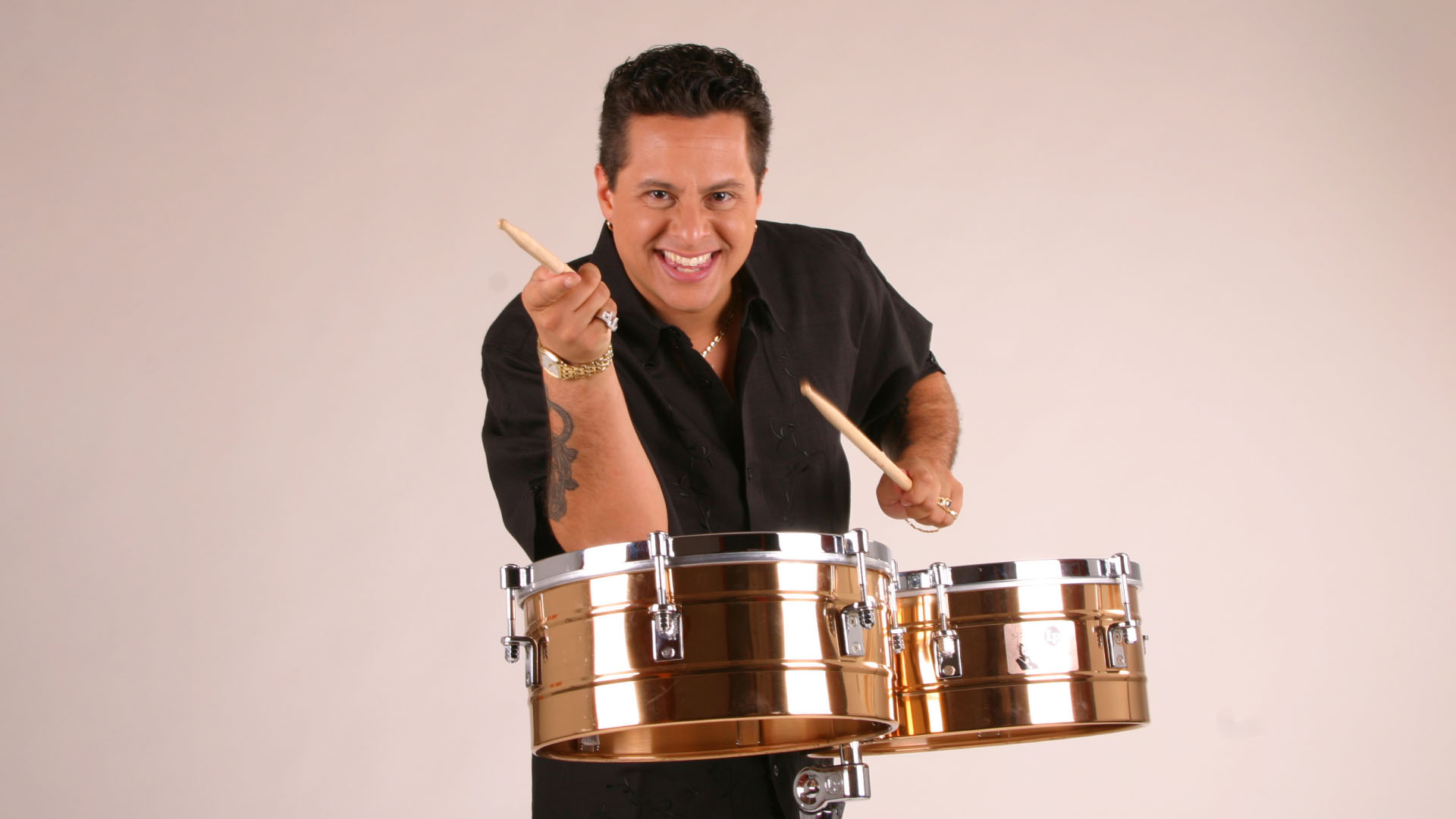 A lively percussionist is captured mid-action, enthusiastically playing bronze timbales with drumsticks. The musician's dynamic pose conveys a sense of passion and energy. The clean, neutral background focuses attention on the performer and the shiny, metallic drums.