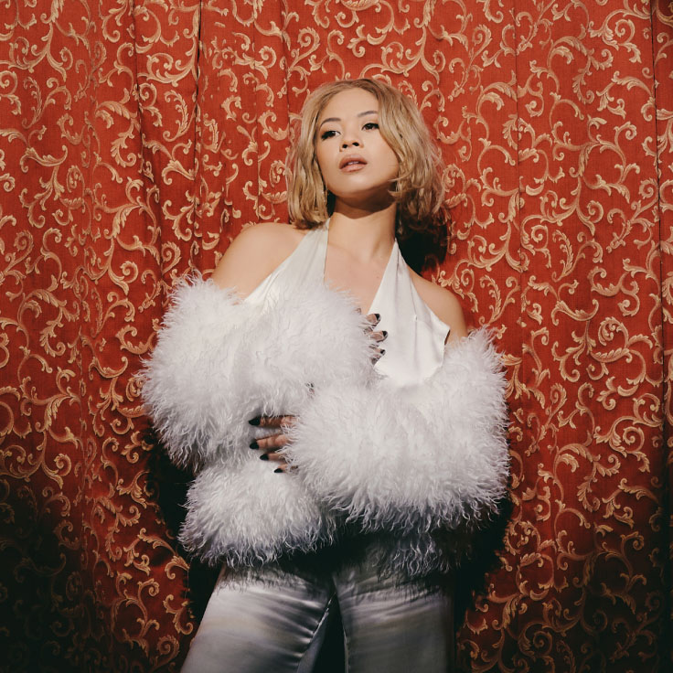 A woman stands confidently against an ornate red and gold patterned backdrop. Her attire features a luxurious white fur coat and shiny satin garments, reminiscent of classic Hollywood glamour. The soft lighting and her poised expression exude sophistication and elegance, capturing the essence of vintage fashion and timeless beauty.