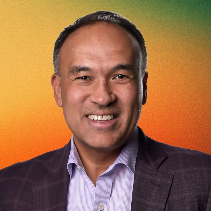 Mark Tatum smiling in a dark blazer and light purple shirt against an orange-to-green gradient background.