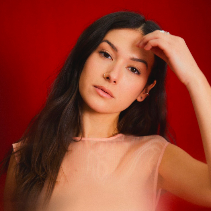 A woman of black hair with her face leaning to the left and her left hand touching her face, dressed in a pink lace blouse