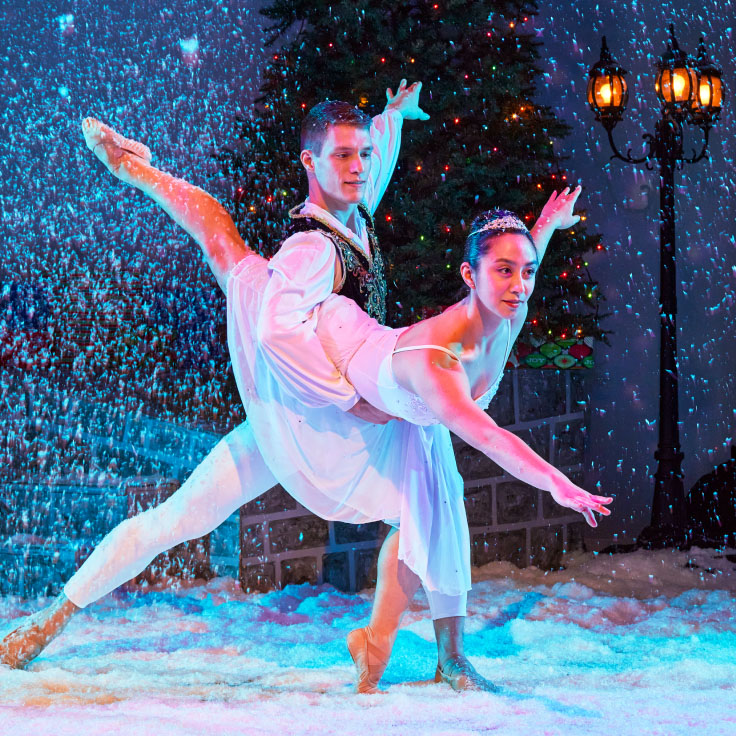 A ballet duo performs gracefully amidst a winter-themed stage, with snow gently falling around them. The dancers are elegantly poised, capturing a moment of artistry and balance. The backdrop features festive decorations, including a beautifully lit street lamp and evergreen trees, evoking a sense of holiday magic and wonder.
