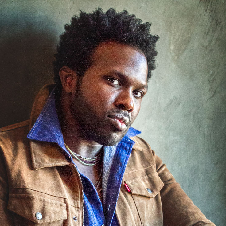 A contemplative man with a short afro hairstyle wears a brown jacket over a blue denim shirt, sitting against a textured gray wall. His thoughtful gaze and casual yet stylish attire evoke a sense of introspection and modern style.