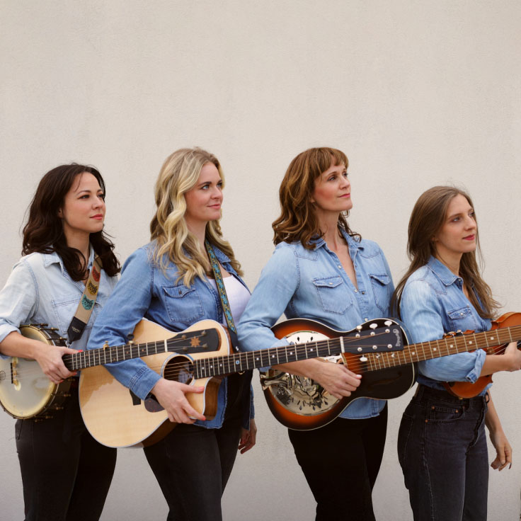Four women musicians stand together, each holding a different string instrument including guitar, banjo, and violin. They are dressed in casual yet stylish attire, with a backdrop of a plain wall, suggesting a setting ready for a casual or intimate performance. Smiling and at ease, the group conveys a sense of camaraderie and joy in music-making.