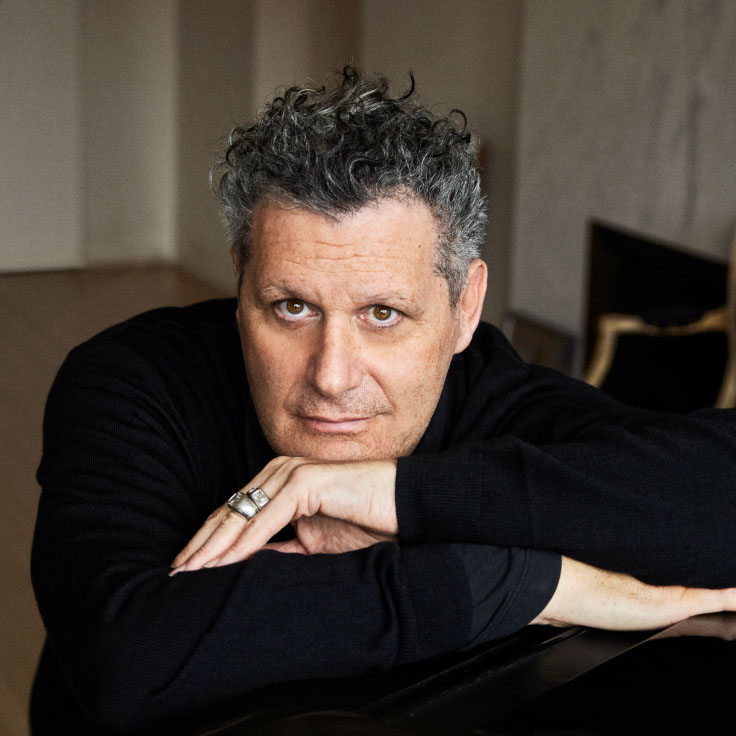 A man with distinctive curly hair is resting his chin on his arms in a dimly lit room. He wears a black shirt and rings, conveying a thoughtful demeanor. The background includes subtle interior details such as a fireplace and a muted color scheme, enhancing the intimate and contemplative atmosphere.