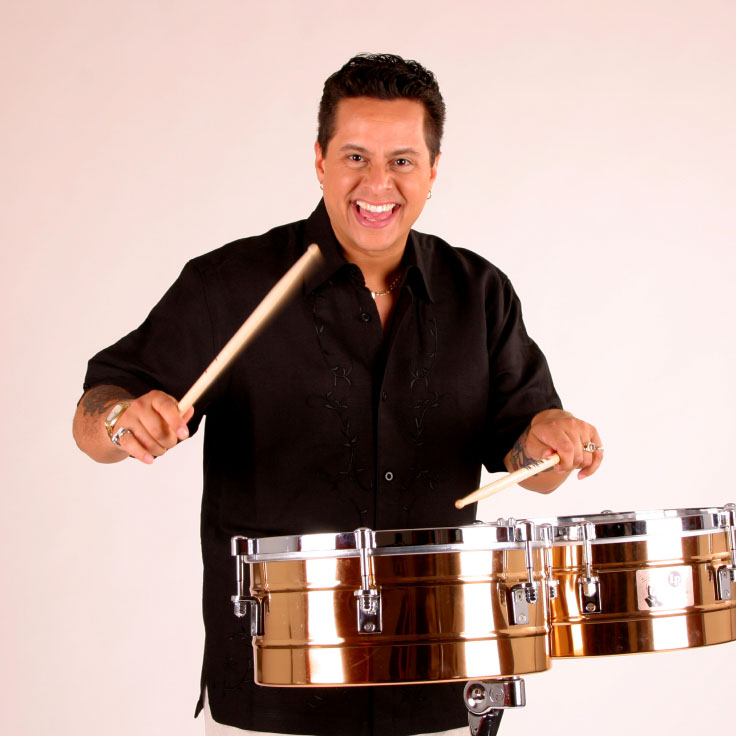 A lively percussionist is captured mid-action, enthusiastically playing bronze timbales with drumsticks. The musician's dynamic pose conveys a sense of passion and energy. The clean, neutral background focuses attention on the performer and the shiny, metallic drums.