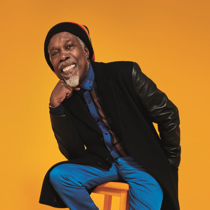 A joyful man wearing a stylish outfit combines vibrant colors and textures, creating a striking contrast against a bright orange background. His attire includes a black coat with leather sleeves, a blue shirt, and blue pants, along with a colorful beanie, embodying warmth and comfort