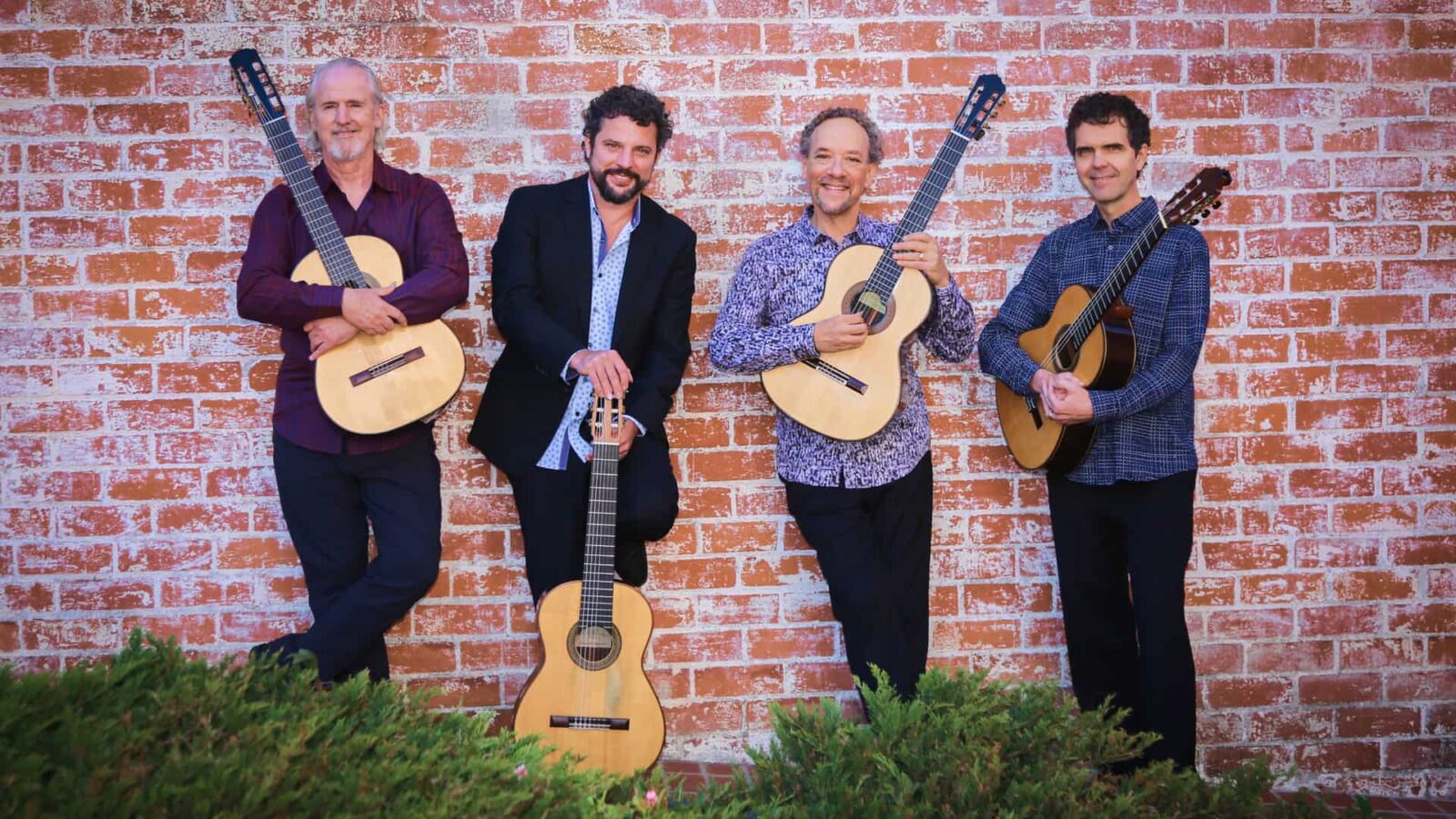 Four musicians stand against a brick wall, each holding an acoustic guitar. They are casually dressed and smiling, conveying a sense of camaraderie and passion for music.