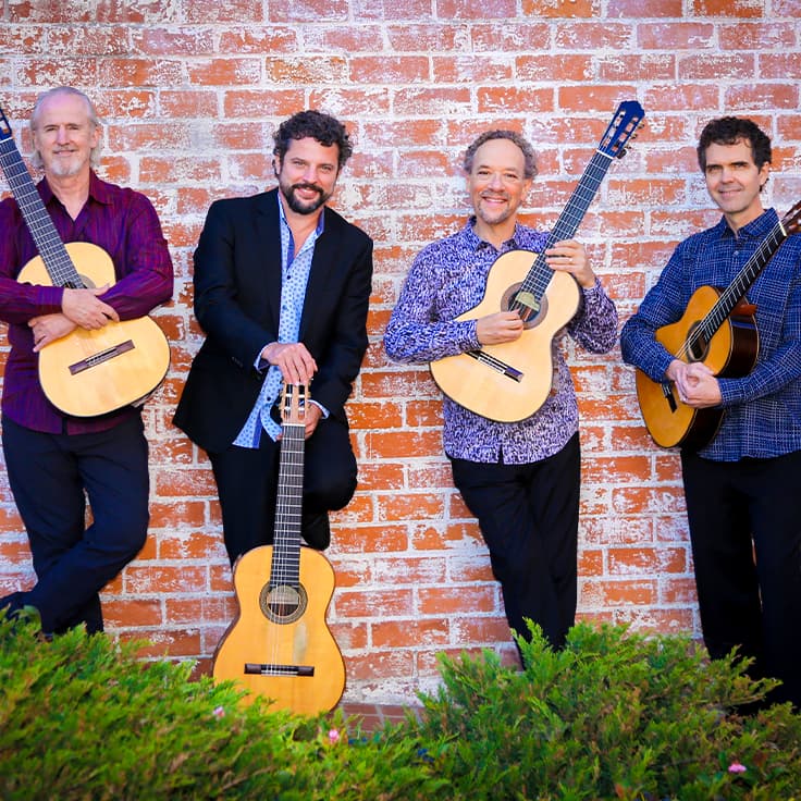 Four guitarists pose casually with their acoustic guitars in front of a red brick wall. They each wear colorful casual shirts, conveying a relaxed yet professional vibe. The lush green plants in the foreground add a touch of nature to the urban setting
