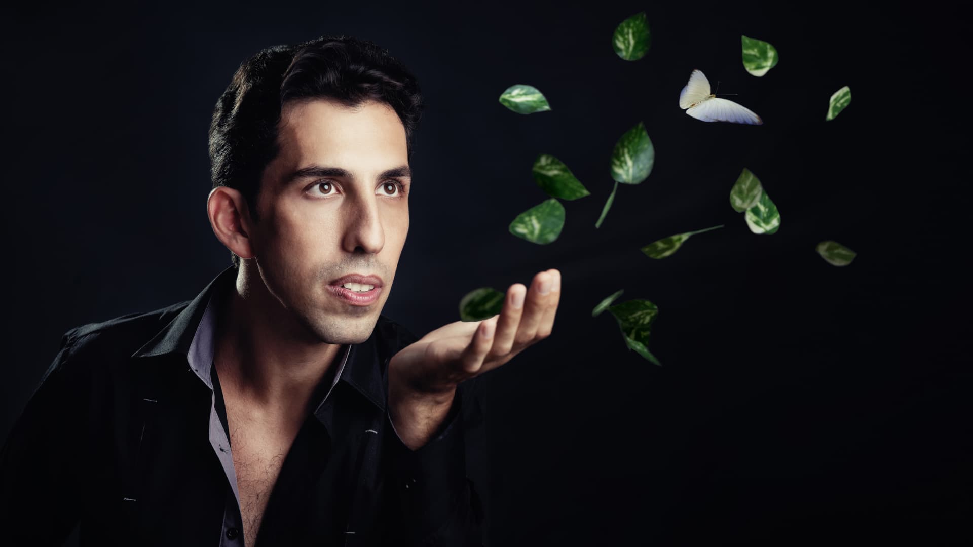 A contemplative man is positioned against a dark background, his hand extended as green leaves and a delicate butterfly seem to float above it, creating a surreal and dreamlike atmosphere.