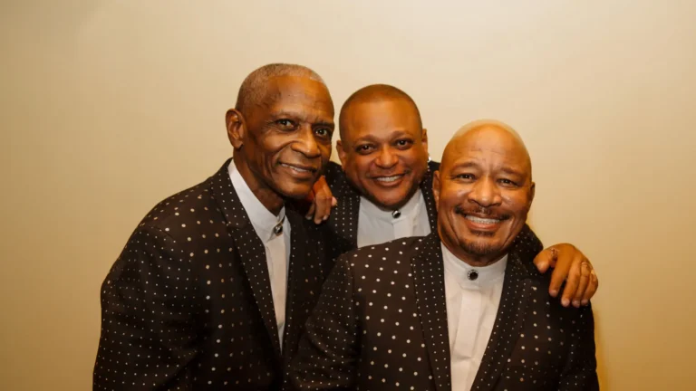 Three men stand closely together, all dressed in matching, polka-dotted black suits with white shirts. Each of them has a warm, engaging smile, suggesting a cheerful and friendly atmosphere. Their coordinating attire and joyful demeanor imply a sense of unity and camaraderie, possibly representing a musical group.